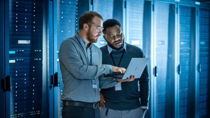 Two data storage pros work on a laptop among servers in a data center.